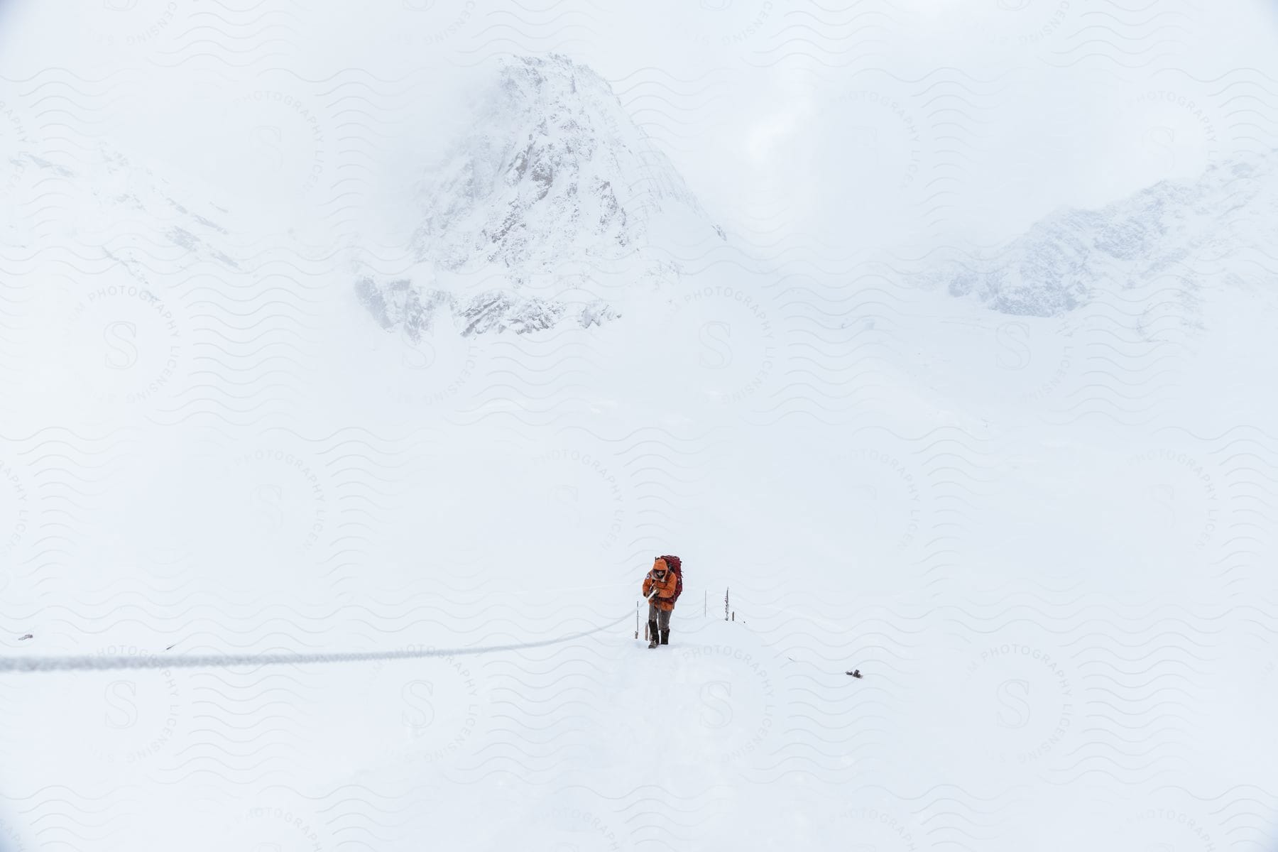 Mountaineer in orange coat and backpack ascending snowy mountain slope using rope