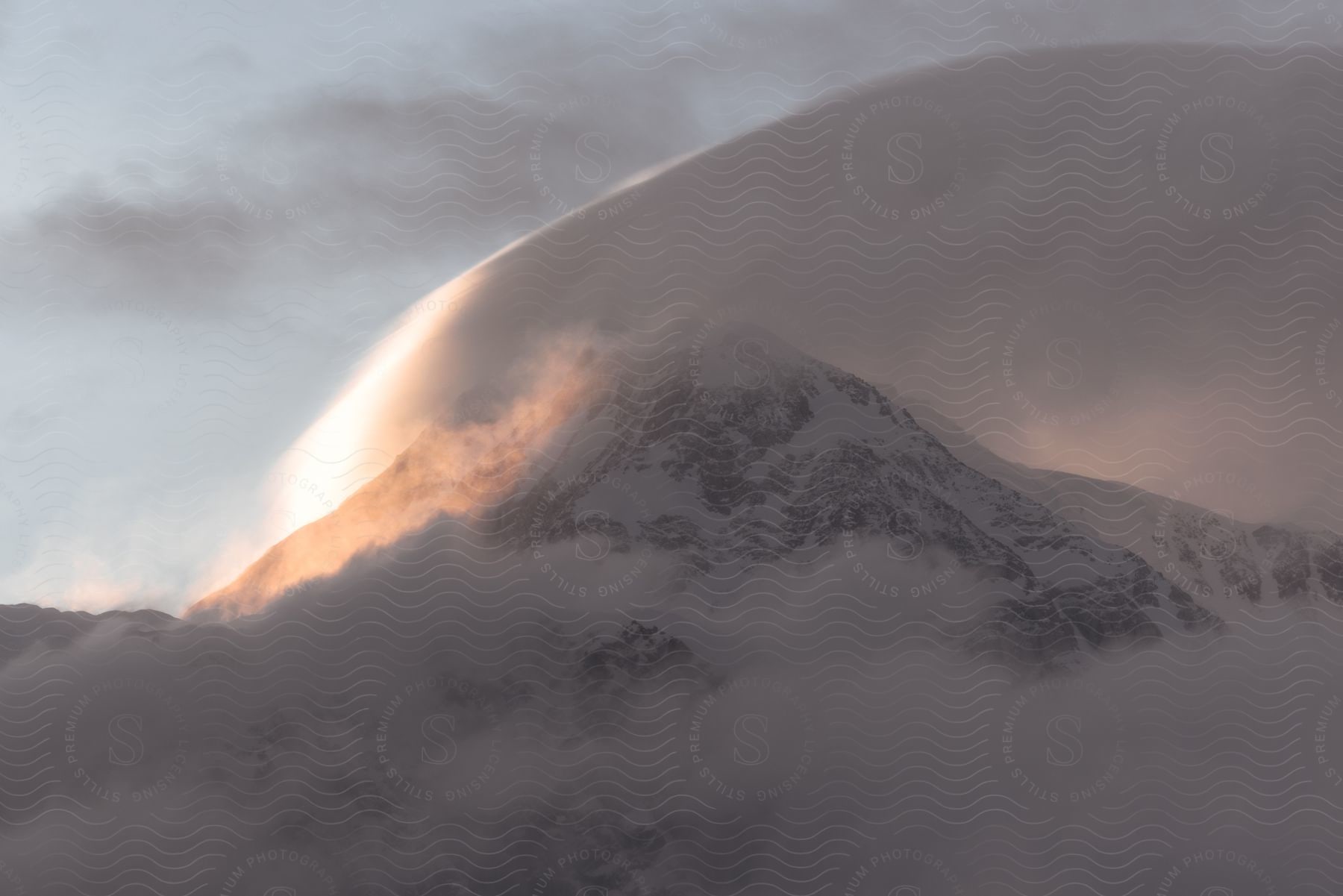 A snow covered mountain with a cloud arching over it illuminated by the sun in a cloudy sky