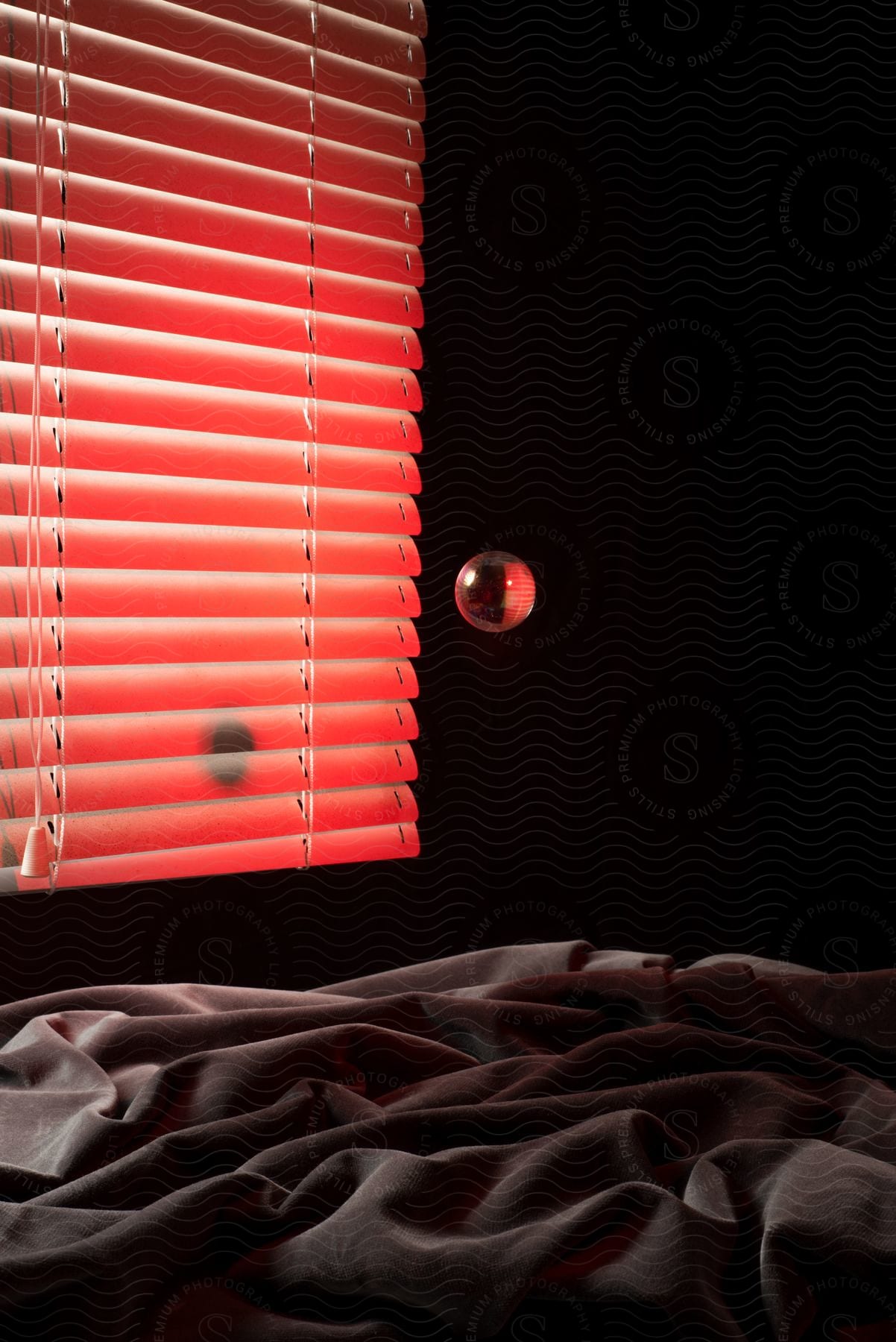 White blinds covering a window with red light shining through and messy bed sheets