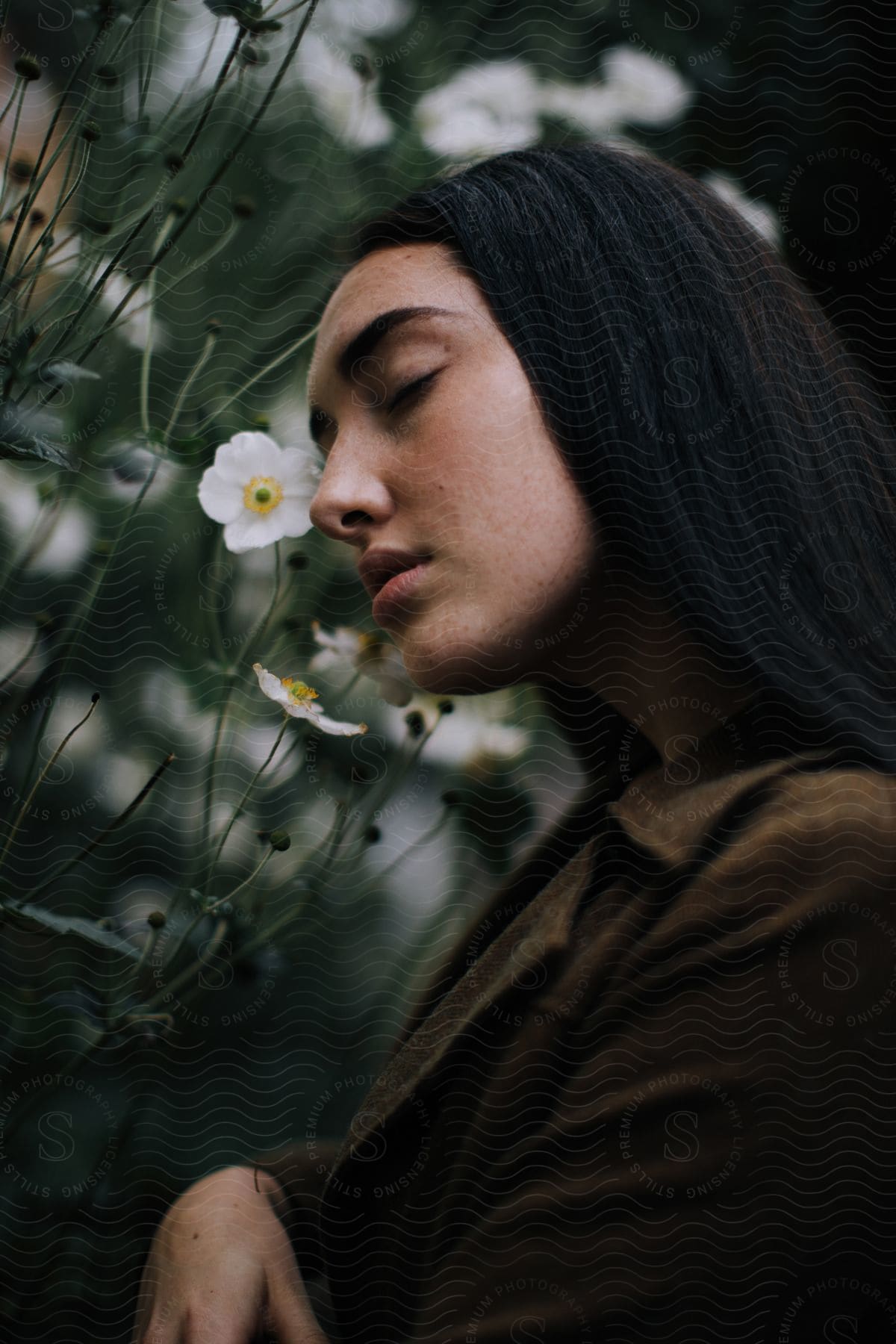 Woman with brown jacket standing next to flowers with her eyes closed