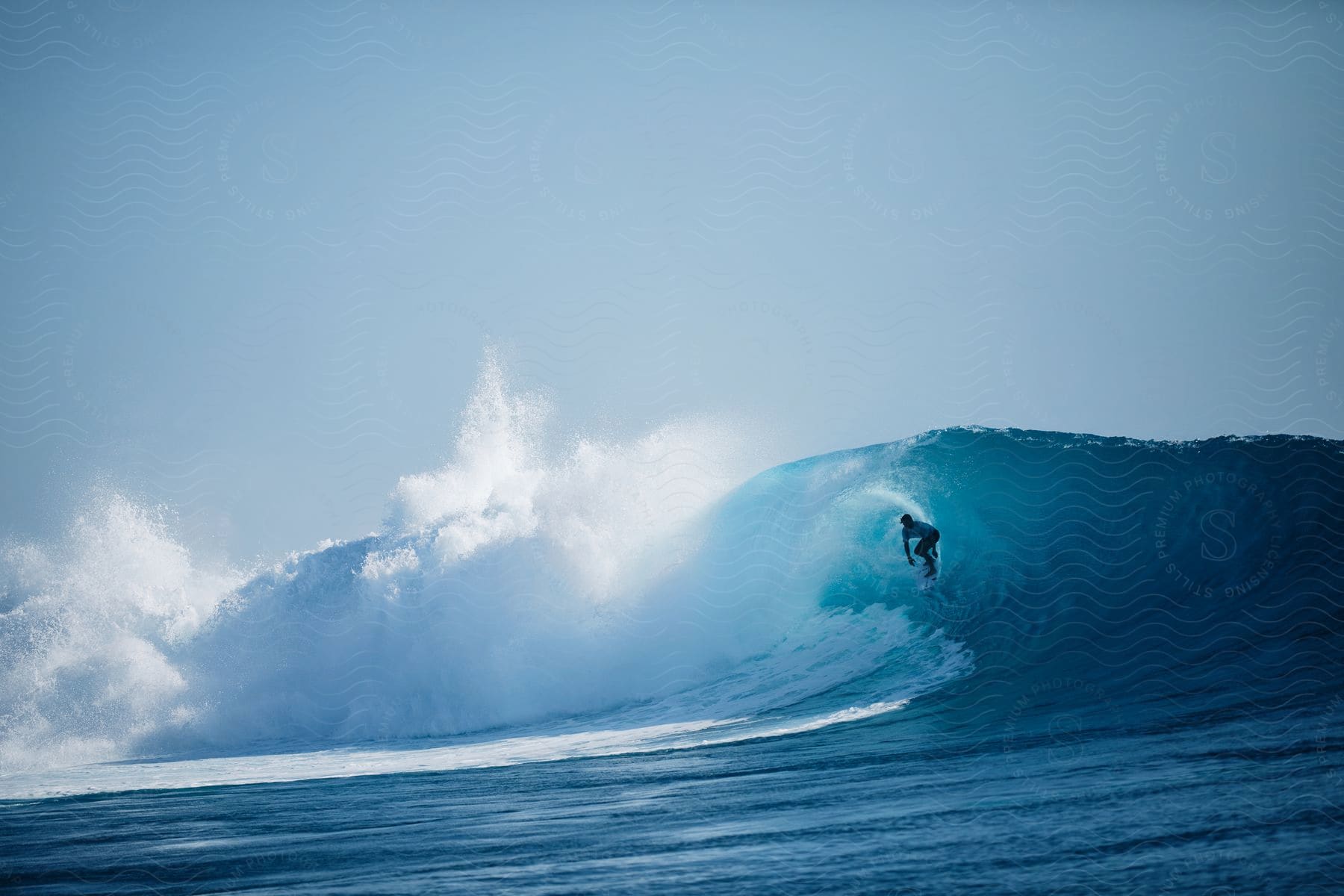 A surfer rides a big blue wave under a clear sky