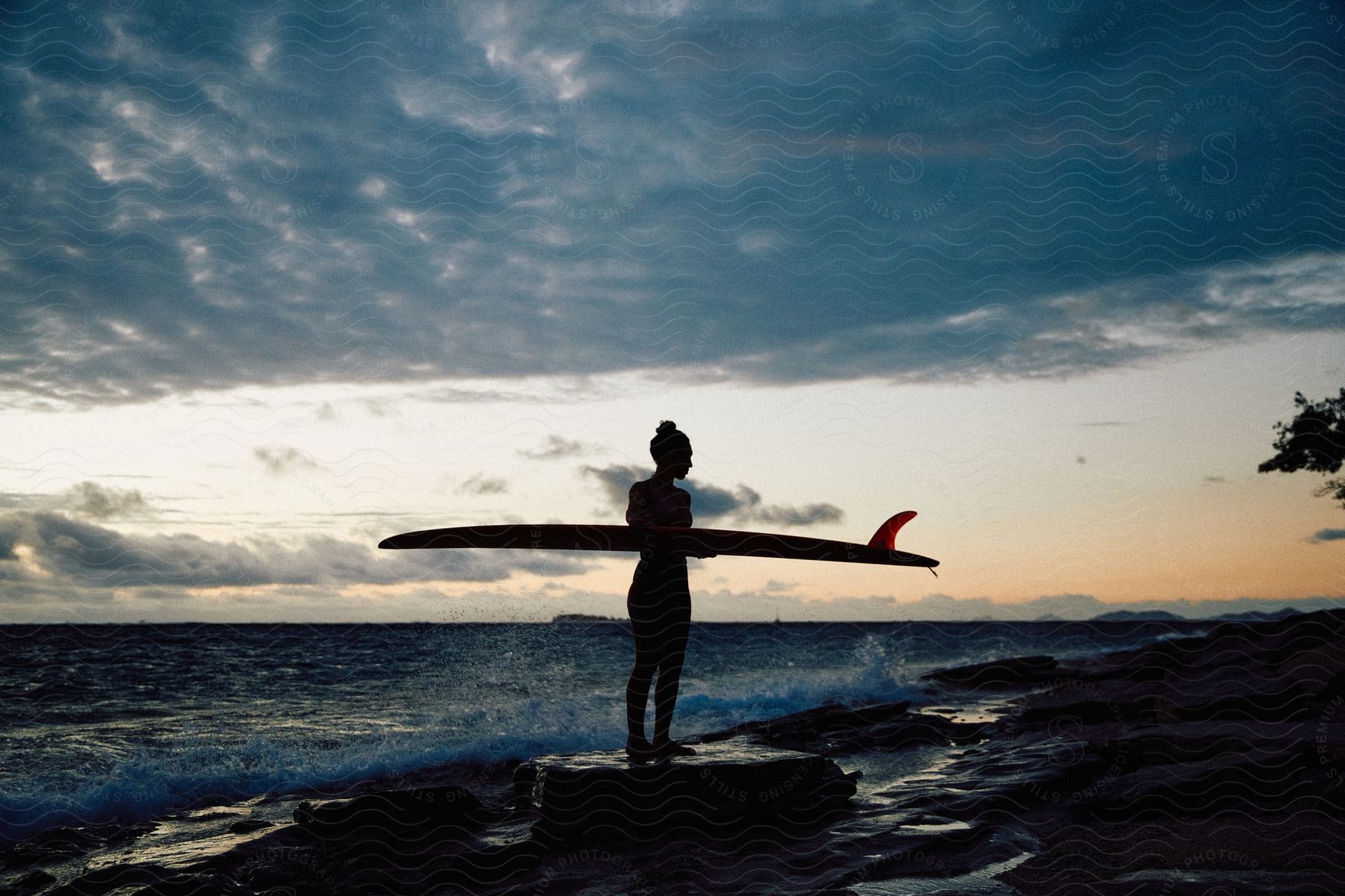 Woman on rock holding surfboard as waves roll into shore under cloudy sky