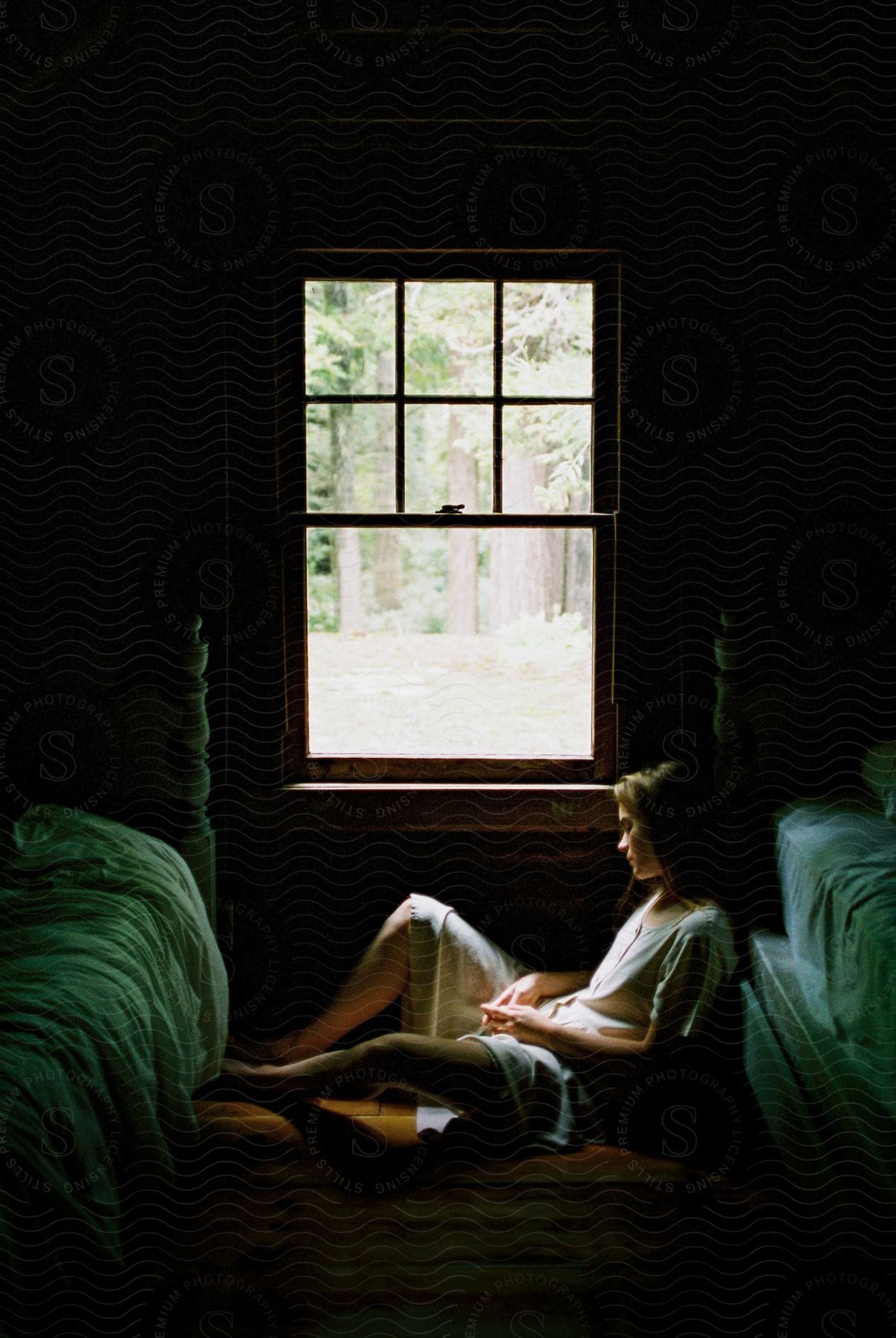 A young blonde woman sitting on the floor next to a window leaning against a bed in a bedroom