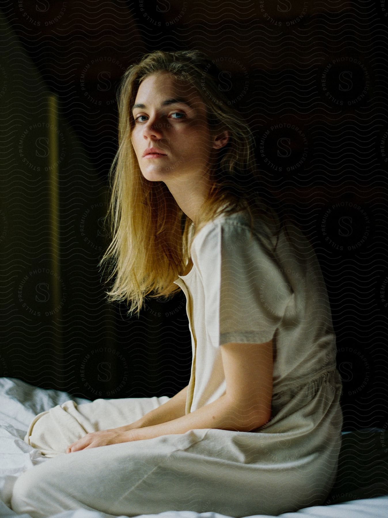 Blonde woman sitting on bed indoors looking straight at the shot