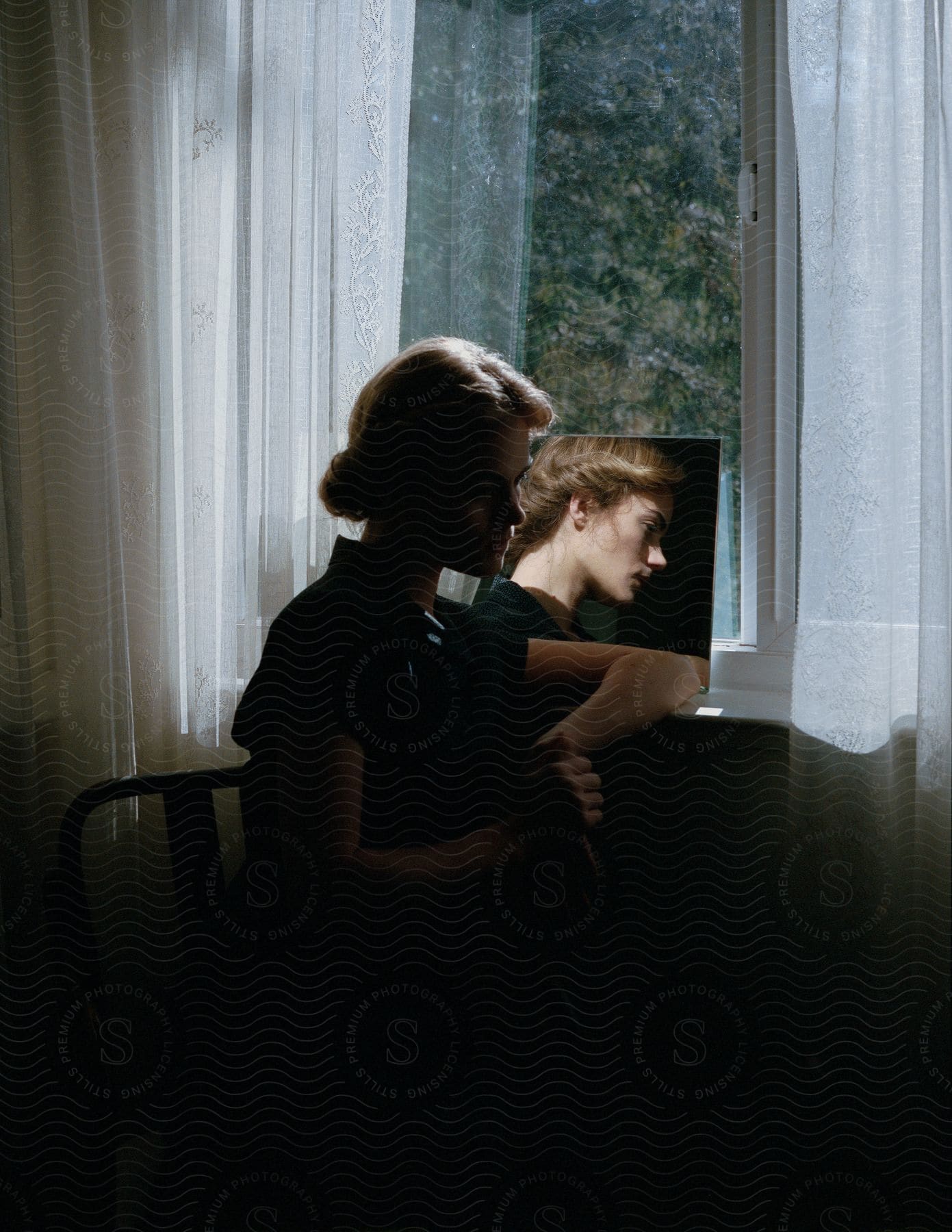 A woman sits and thinks near a window with a mirror leaning against it