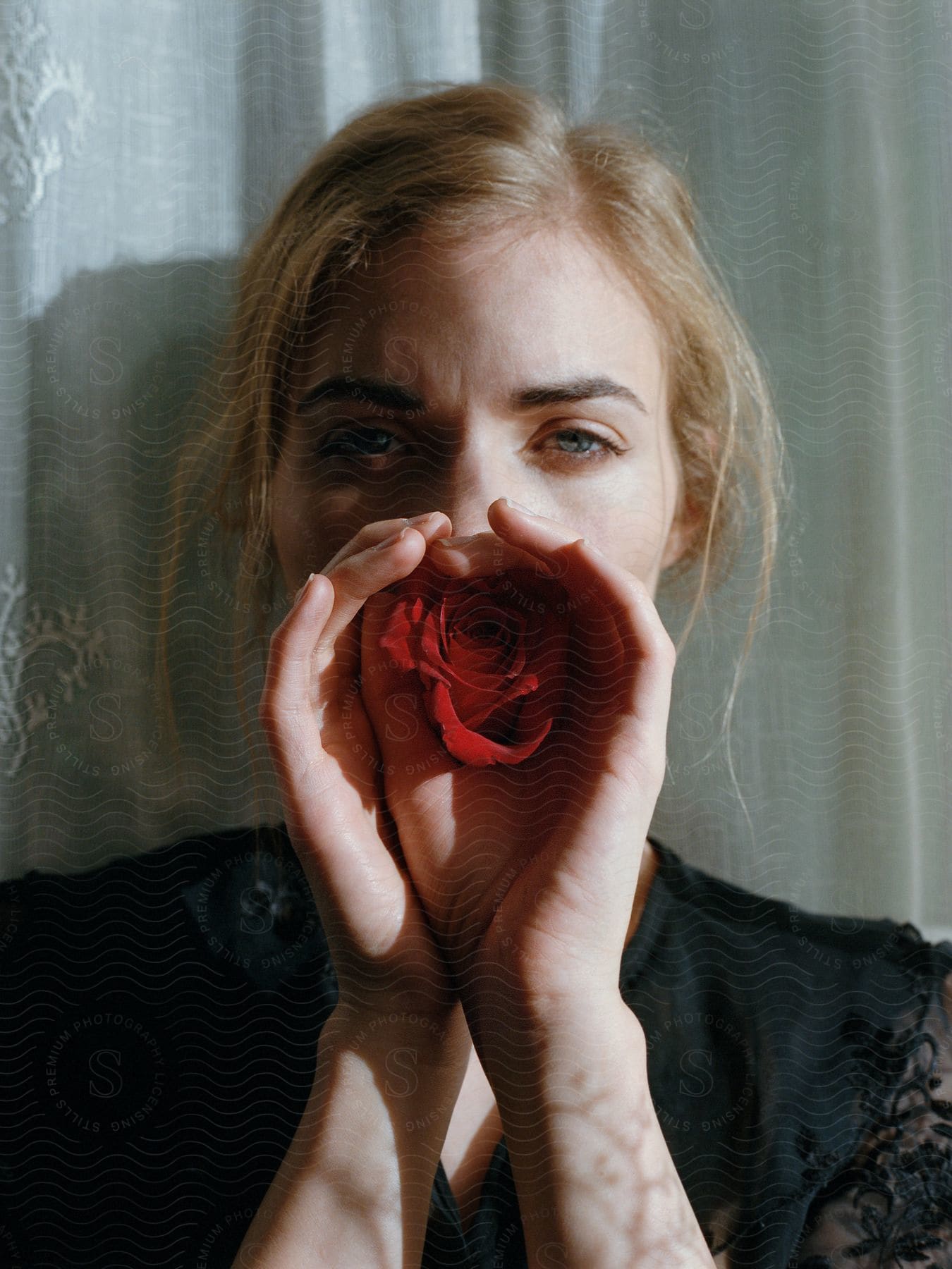 Blonde woman in black attire with hands covering half her face holding a red rose