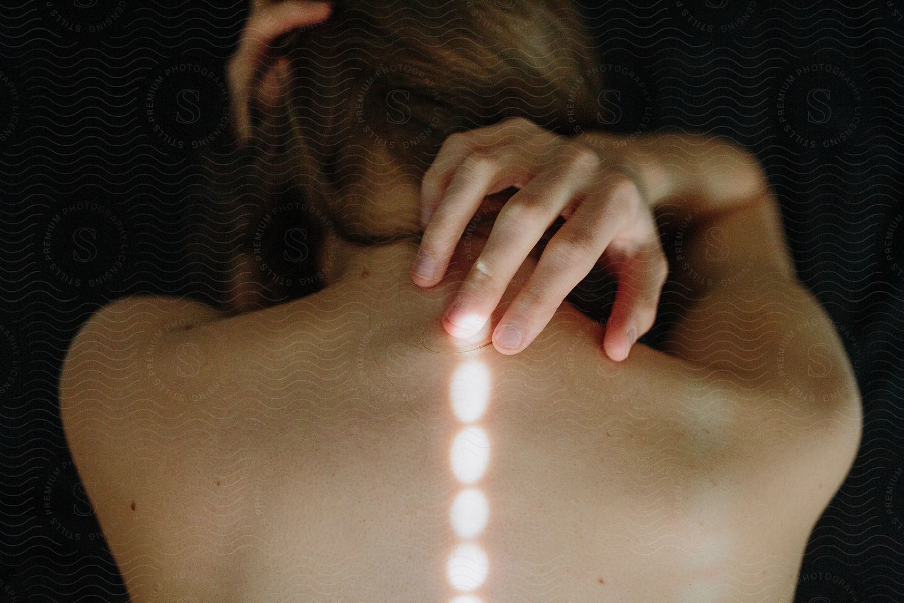 A womans back with her hand on her neck without a shirt illuminated by light