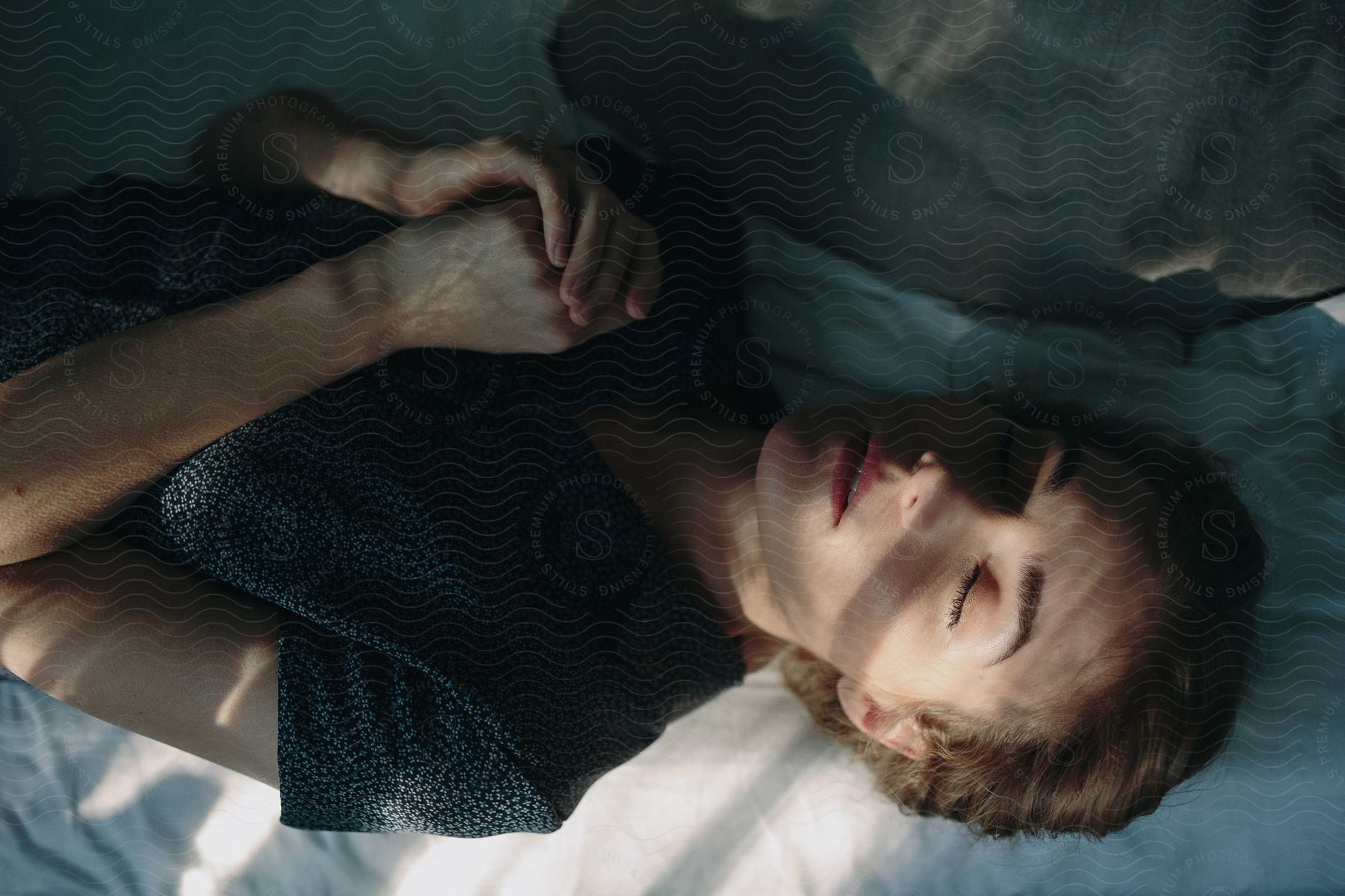 Woman lying on bed with closed eyes and hands together sunlight casting shadows on her face