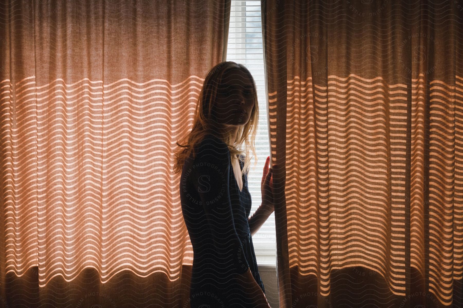 A Young Adult Female Stands Between Curtains In A Dark Room With Sunlight Shining Through The Window Blinds Behind Her