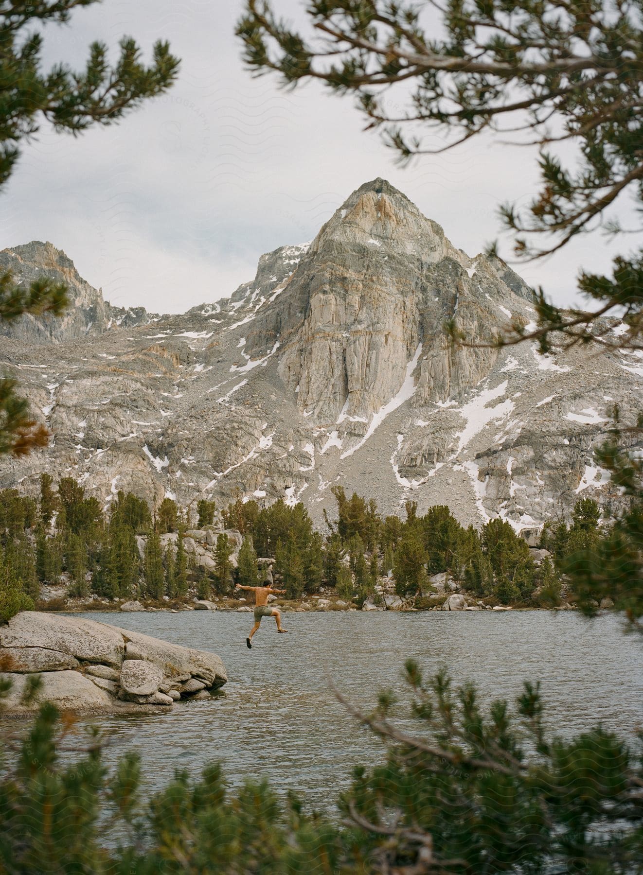 A man jumps into a serene lake surrounded by trees and rocks with a tall mountain in the backdrop