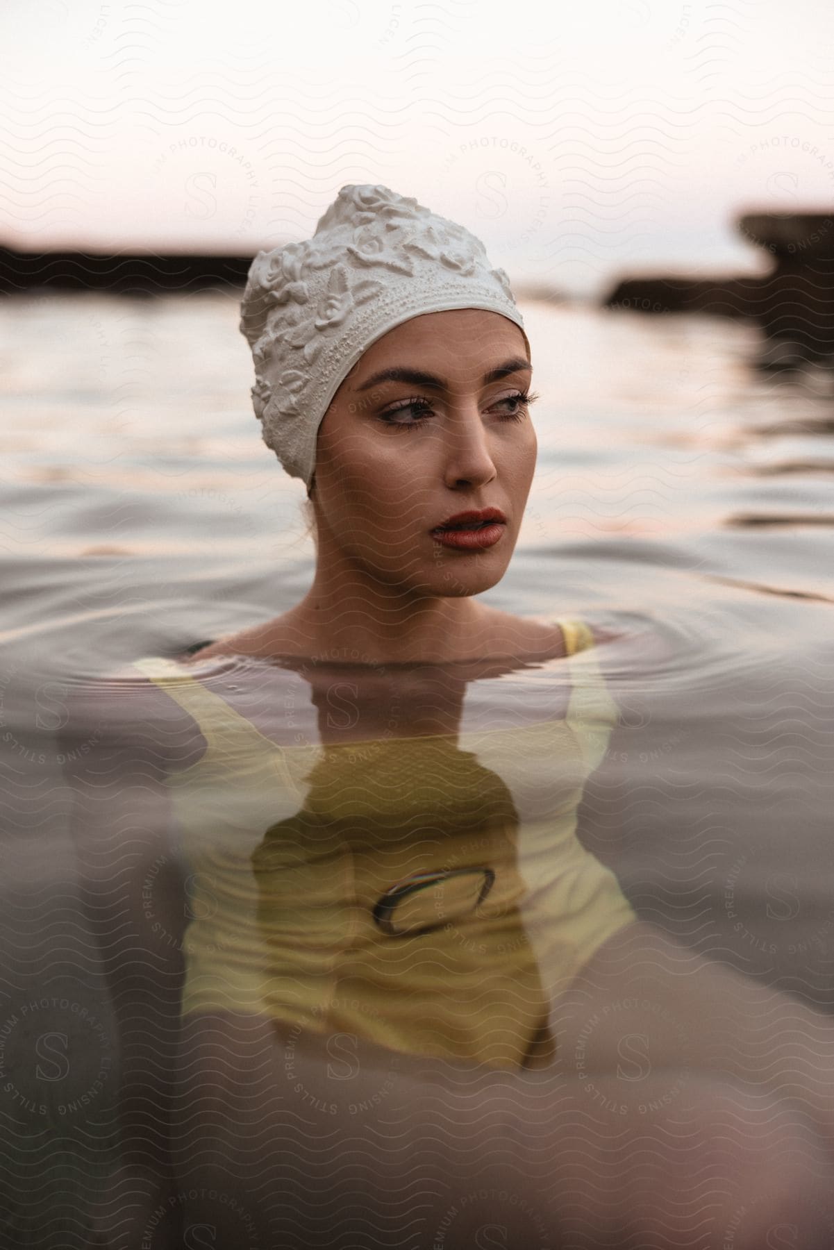 A woman in a yellow swimsuit and white swim cap sitting in clear water looking to the side