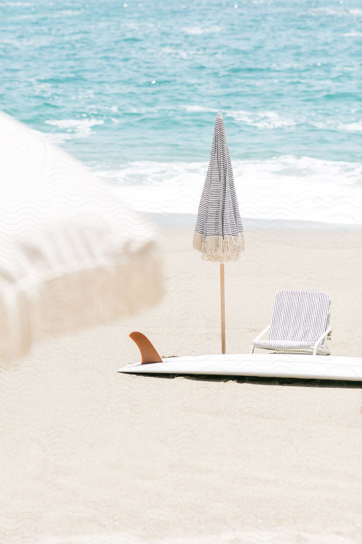 A closed beach umbrella chair and surfboard on an empty sand beach with clear ocean