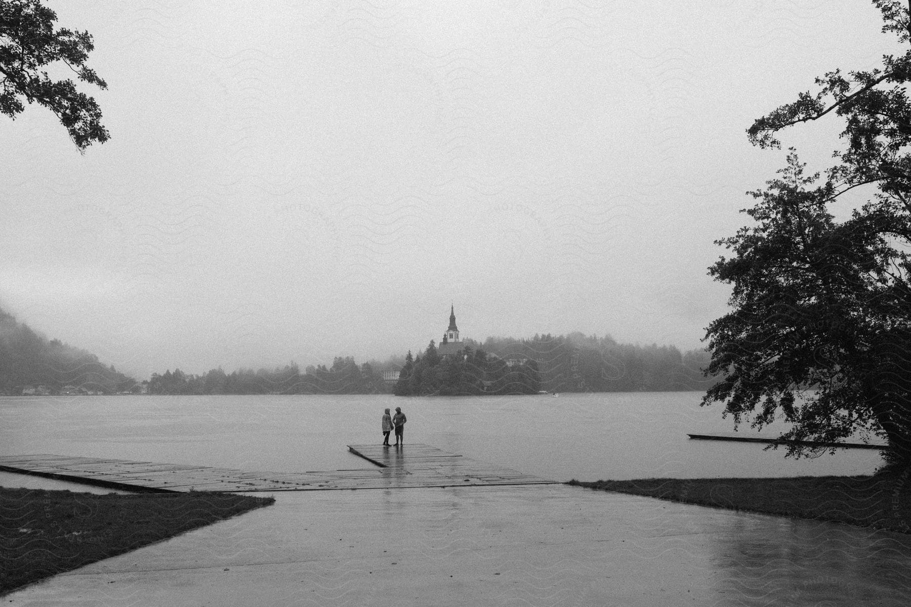 Two people stand on a dock overlooking a lake with a church on an island