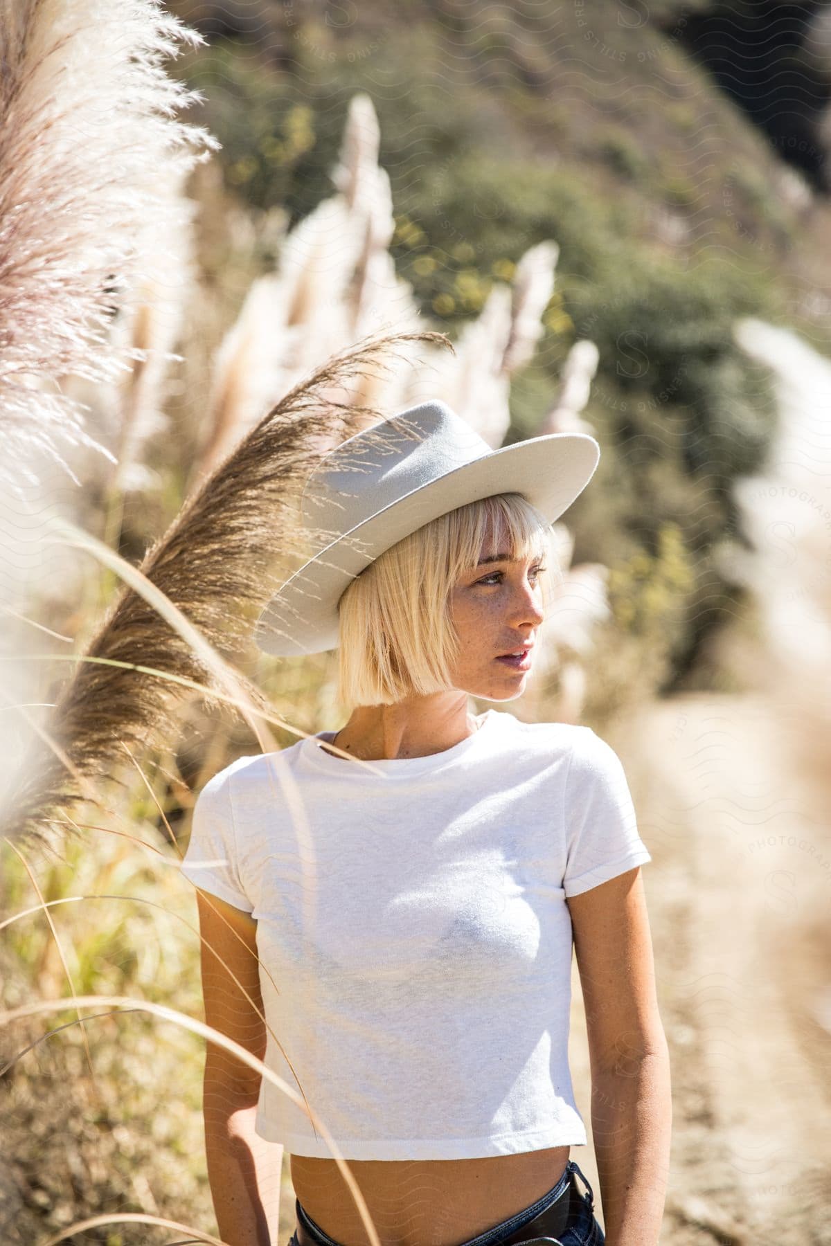 Blonde woman in white hat and tshirt stands outside in the sun near plants and bushes