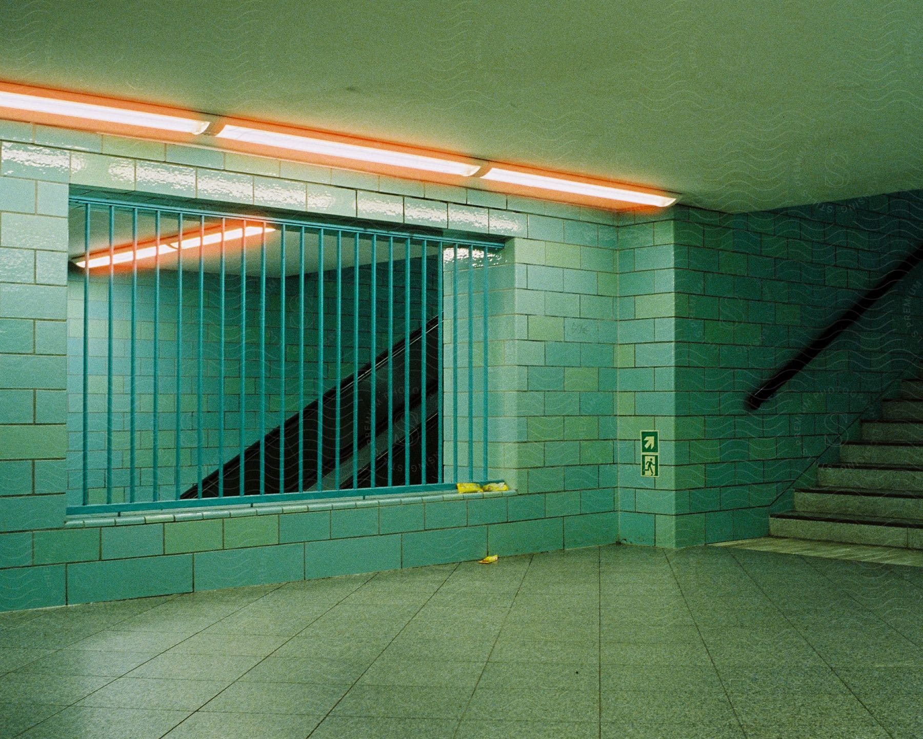 Orange ceiling lights shine down on green tiled walls in an underground tunnel with dark stairways