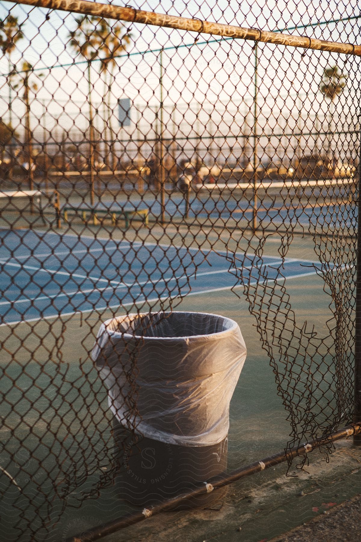 A cloudy day view of a trash can next to a ripped chain link fence surrounding an outdoor tennis court