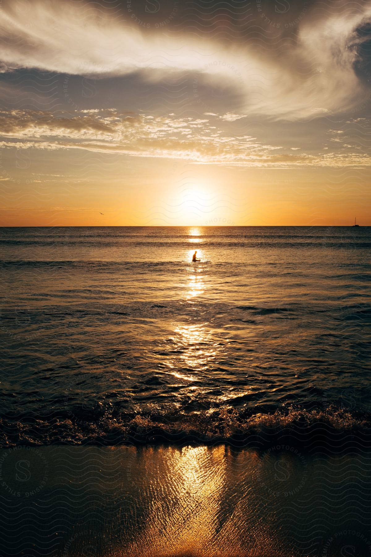 A man sits on the shoreline as the tide comes in in the path of the setting sun