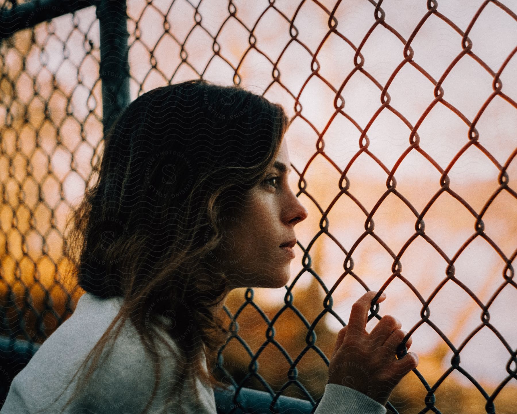 Woman with brown hair and gray sweater looking through chainlink fence at sunset