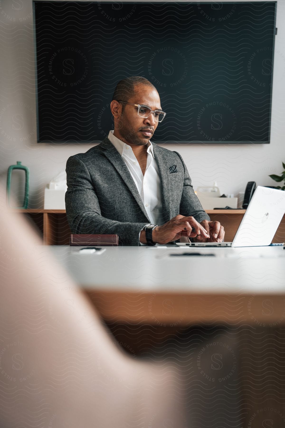 A man wearing a wool dress coat and glasses types on a laptop in an office meeting room