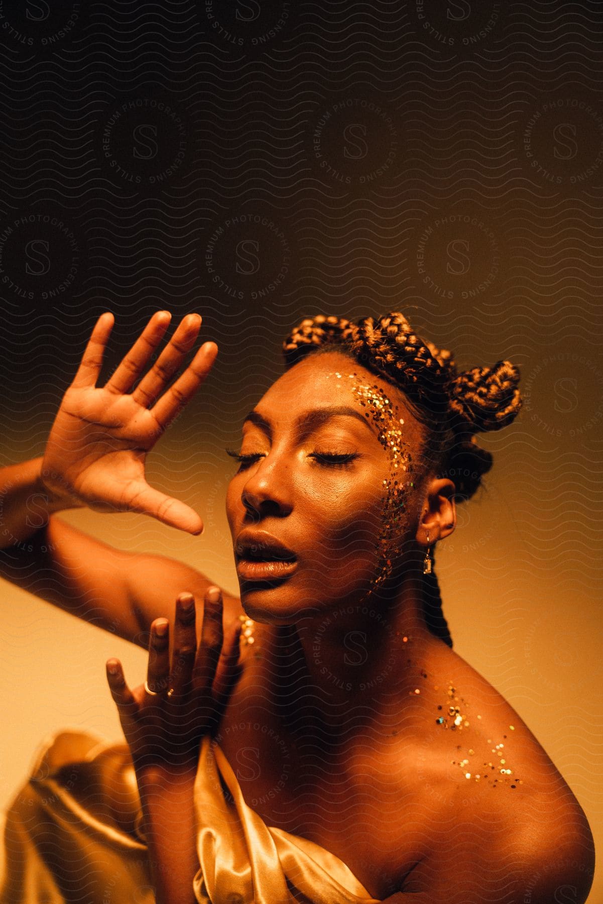 Black girl with raised hands braided hairstyle and golden decorations on her face illuminated by yellow light