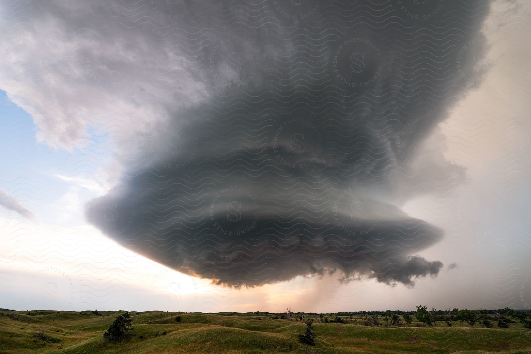 A supercell storm looms over hills and trees as the sun shines in the distance