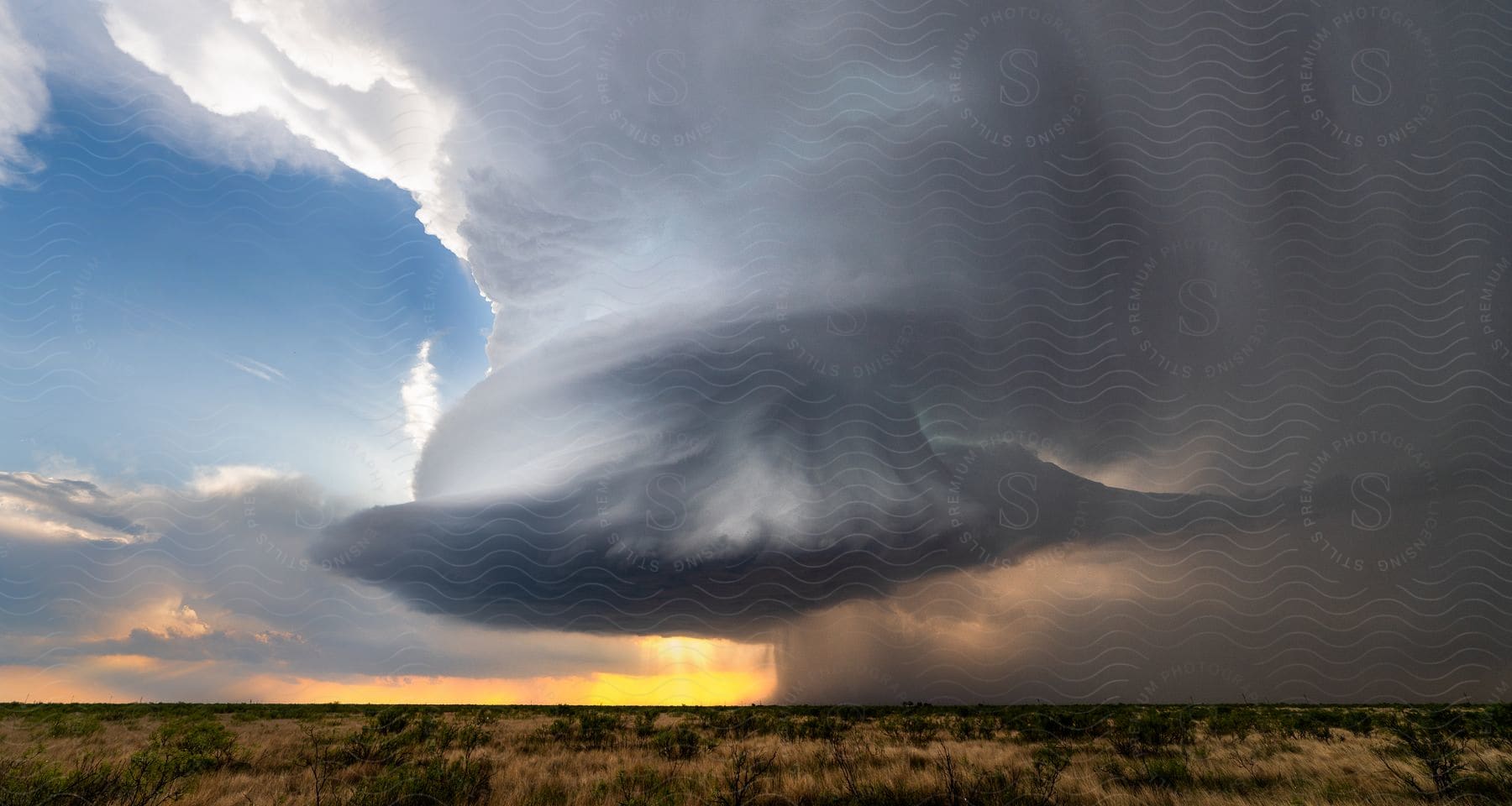 A supercell storm cloud pours rain over rural farmland as the sun shines on the horizon