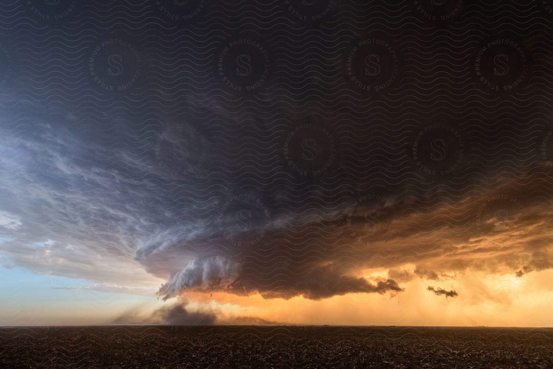 A supercell storm cloud hangs over farmland in an orange sky