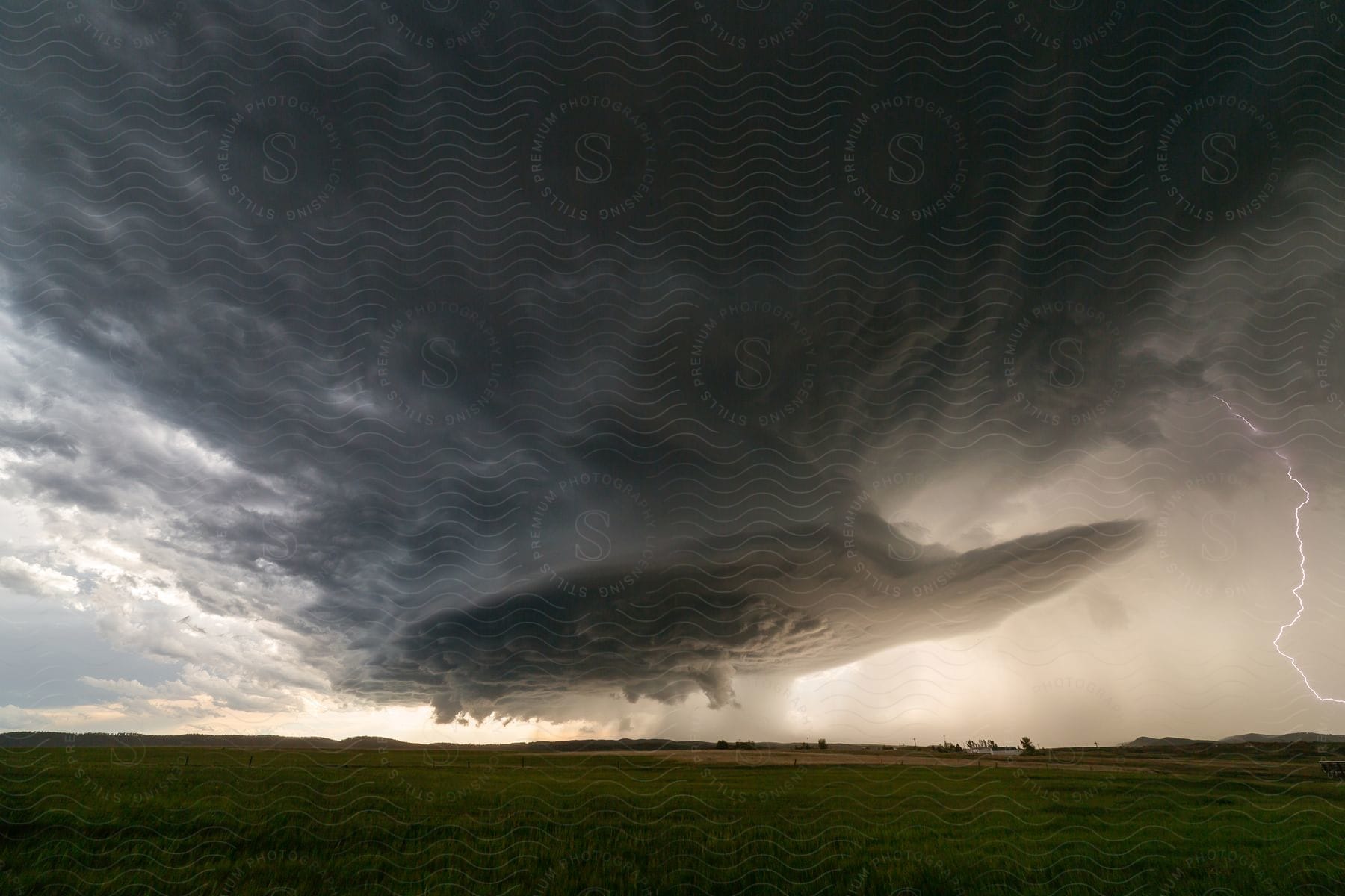 A dark supercell storm pours rain over rural land as lightning emerges from the cloud