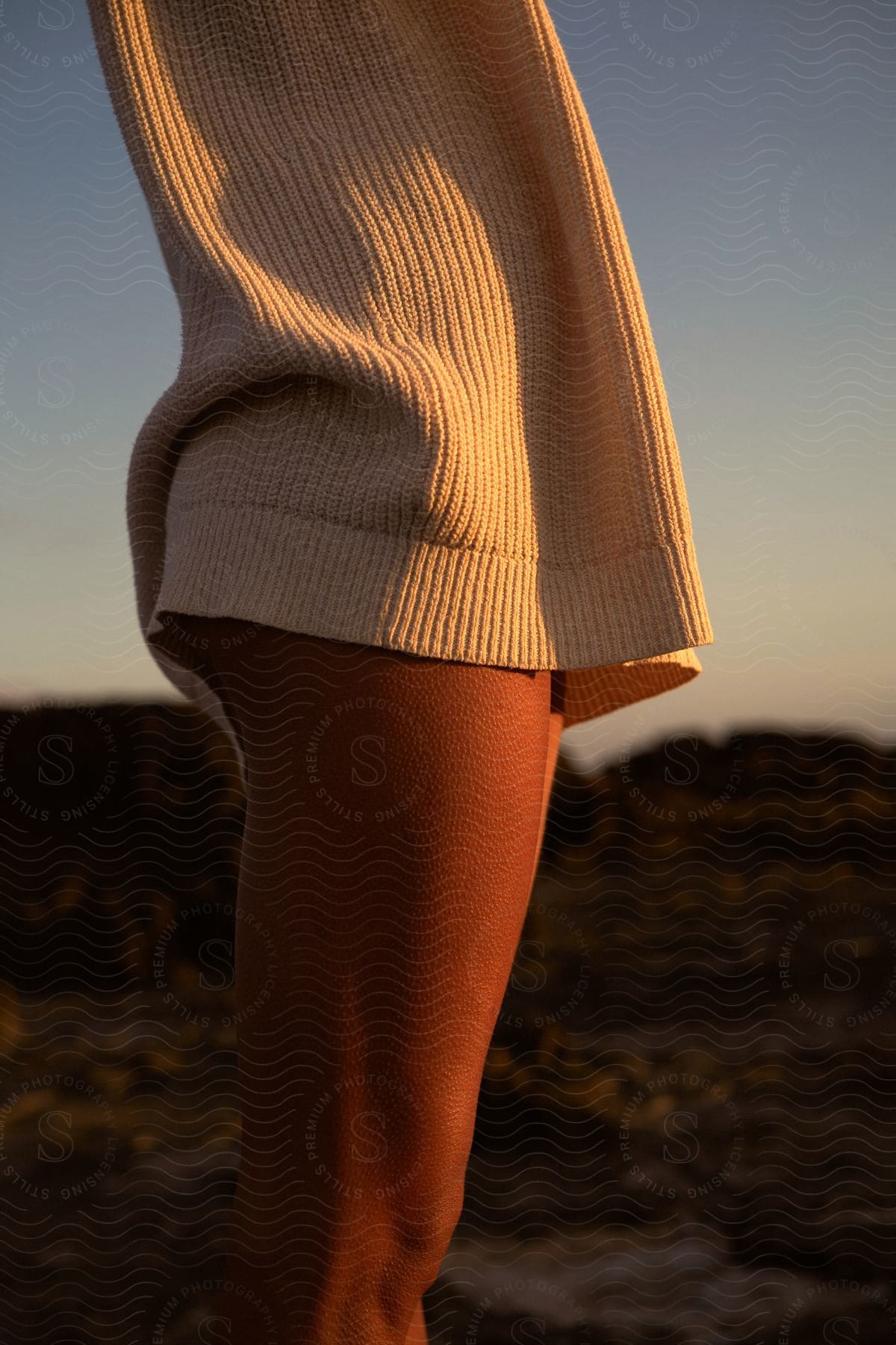 Woman wearing a white sweater and bikini stands on the coast watching the sunset