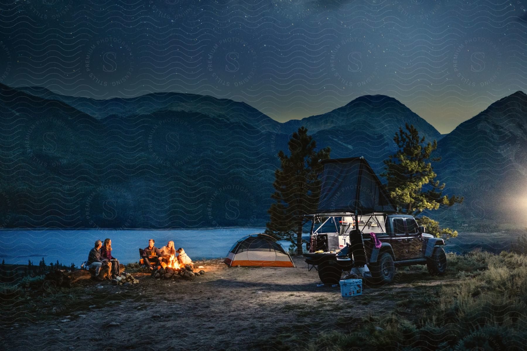 Two couples with their dogs sitting by a campfire at the lake with mountains behind