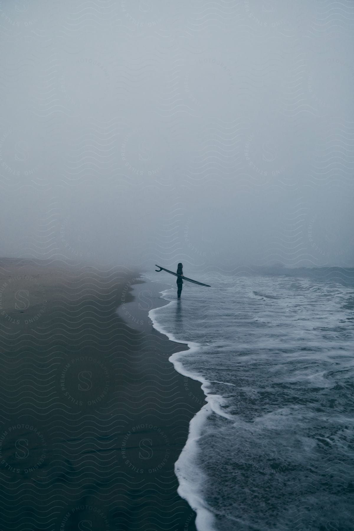 Woman holding surfboard in water on beach with waves under foggy sky