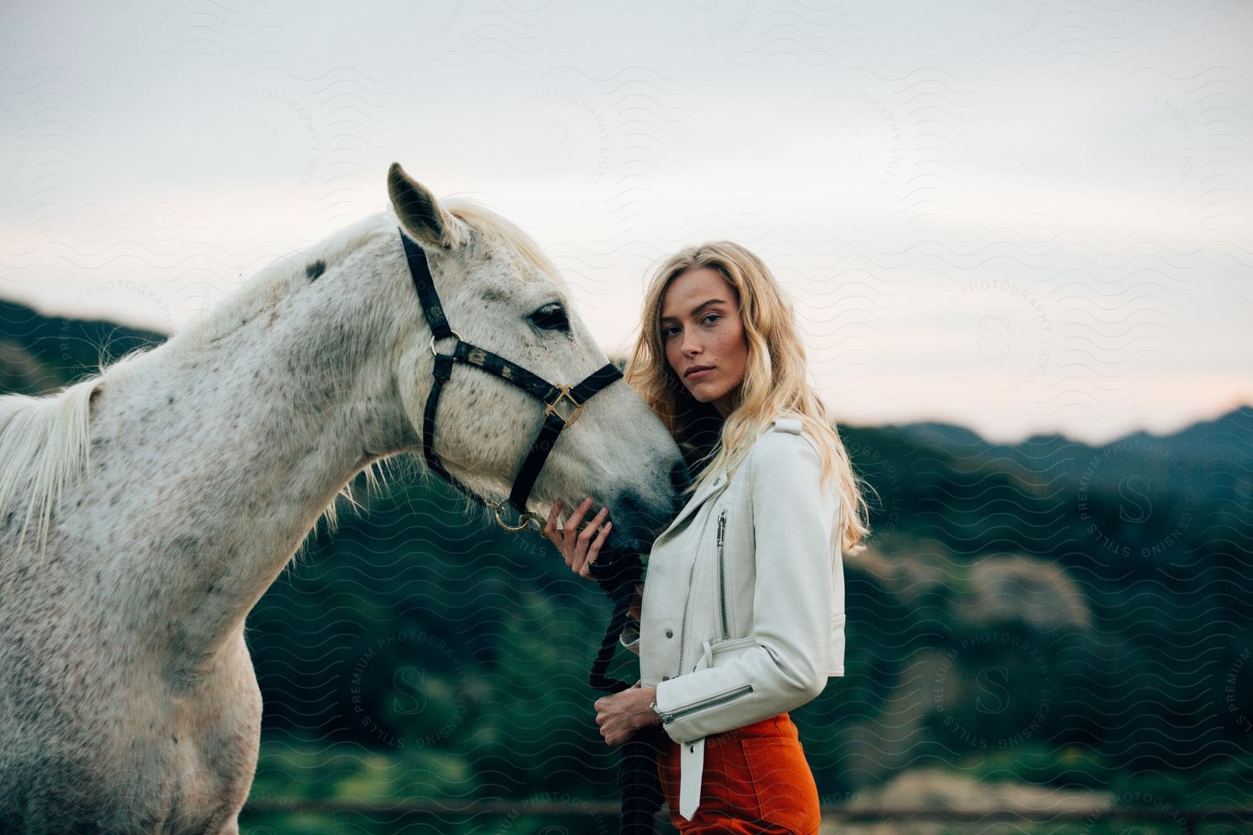 Blonde woman holding the head of a horse in the countryside looking at the camera