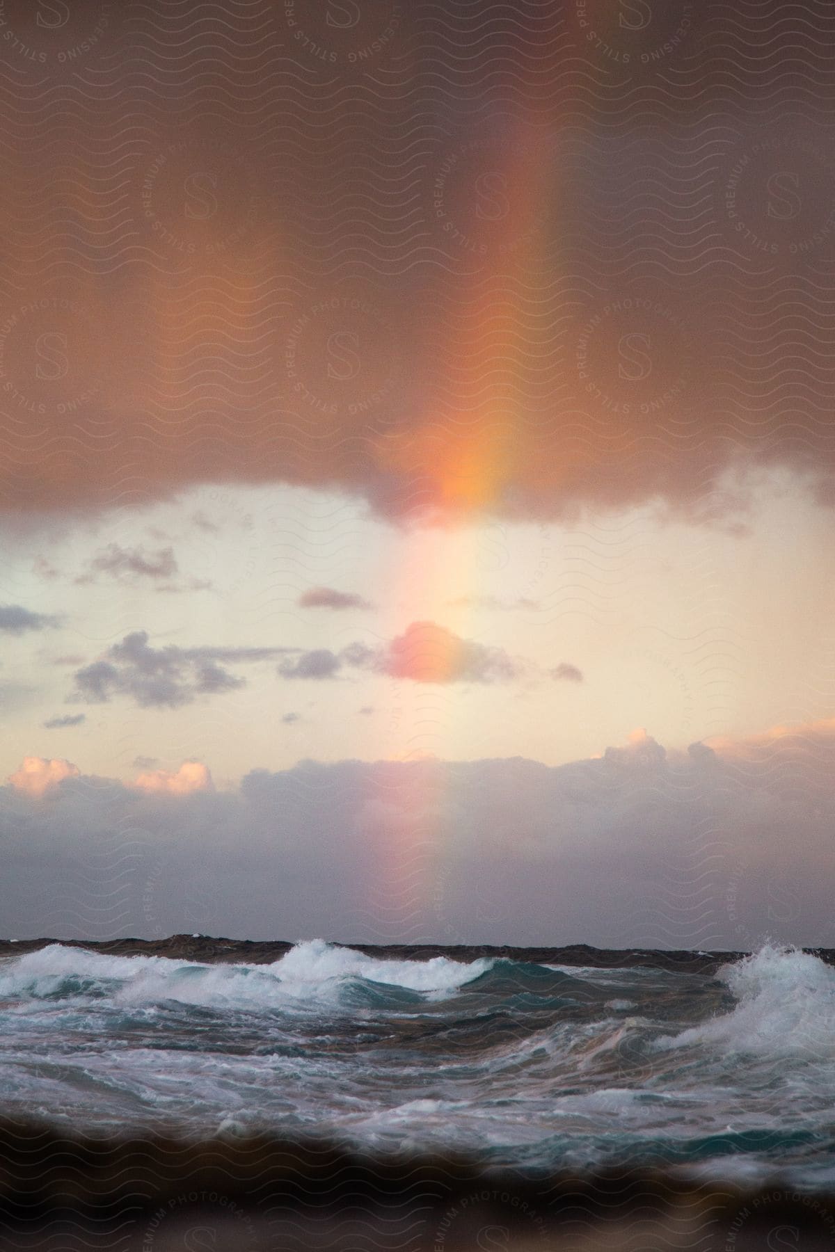 A rainbow extends from a purple cloud to the ocean water below as waves roll in