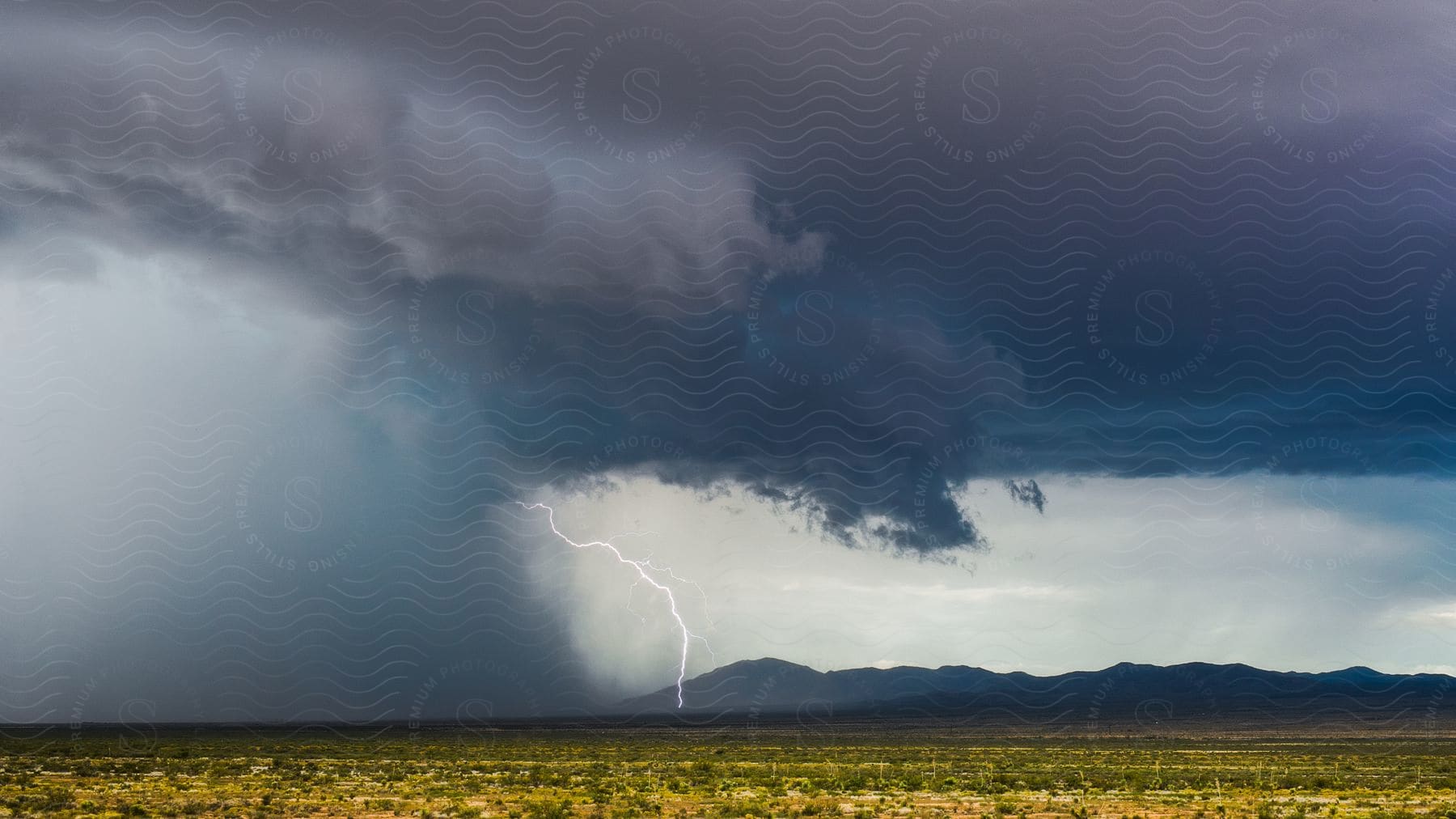 Lightning strikes from a supercell storm cloud moving over farmland with mountains in the distance