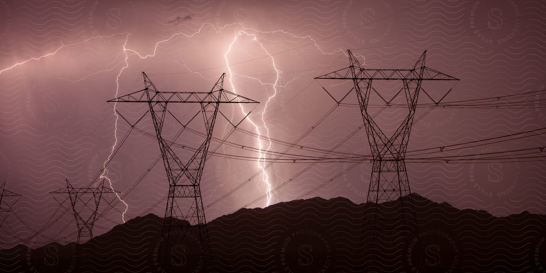 Cables stretch across utility towers as lightning illuminates the stormy night sky over mountains