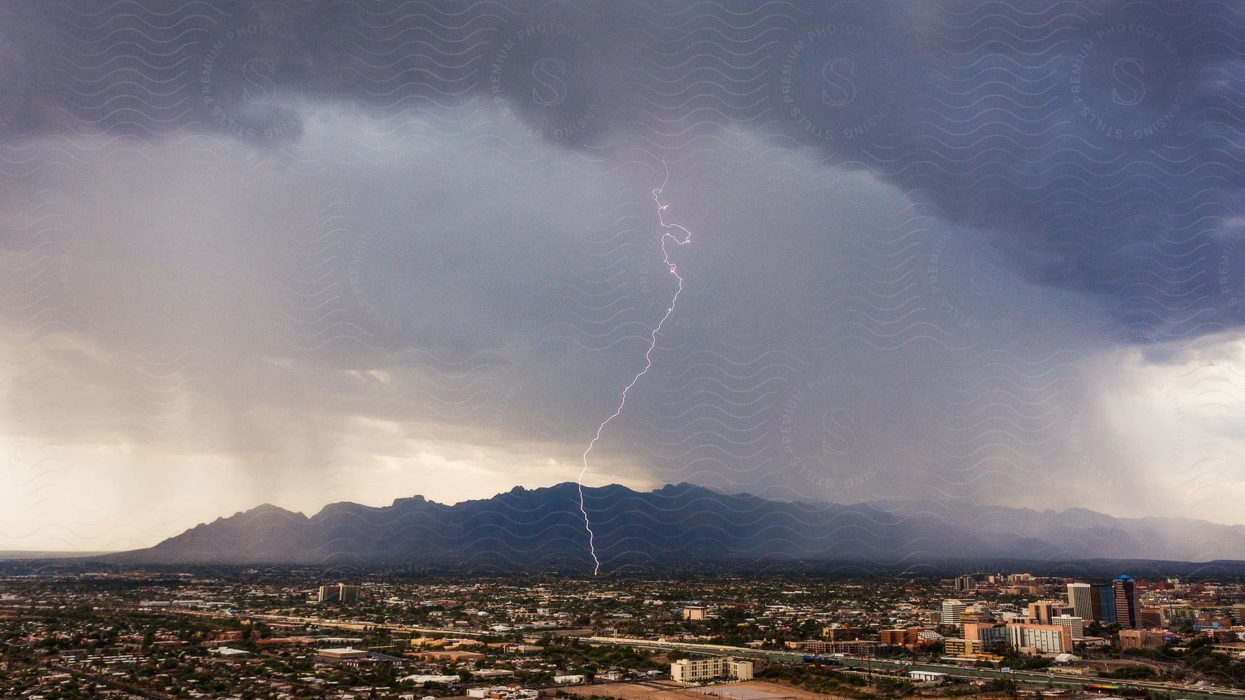 Stock photo of lightning bolt strikes near a town under thick clouds with mountains and bright sky in the background at dusk