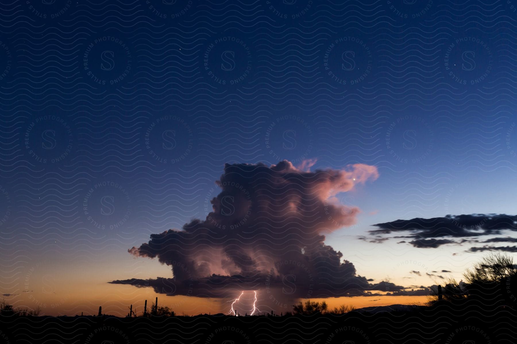 Lightning strikes from a dark supercell storm as it pours rain over the desert at sunset