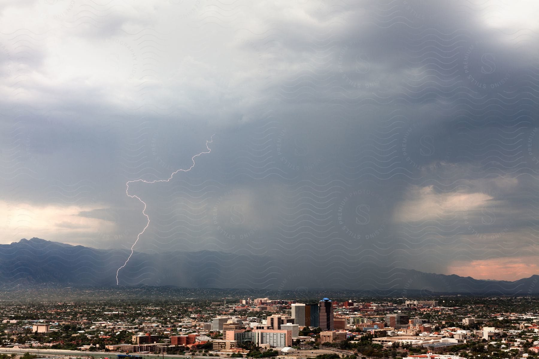 Lightning strikes from above as storm clouds pour rain over a city with mountains in the distance