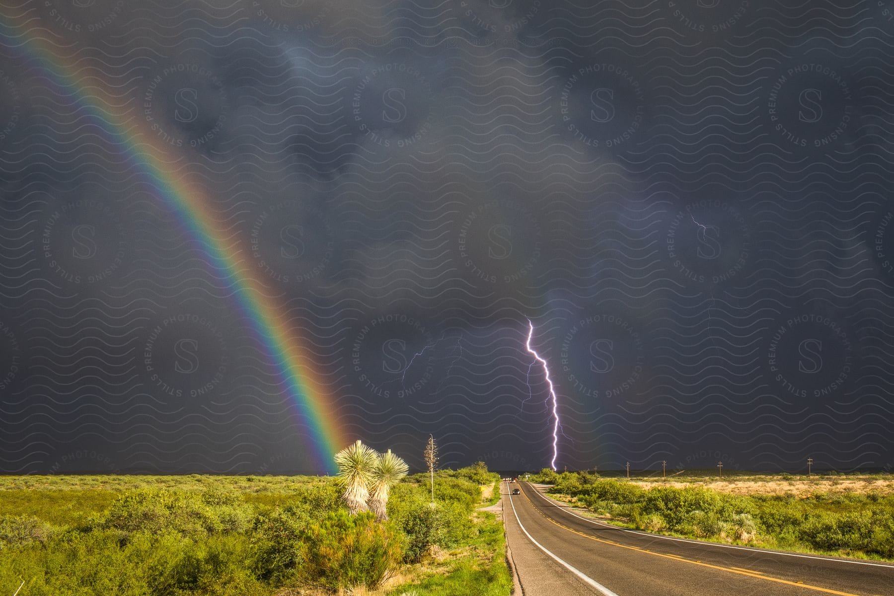 An automobile drives on a road as lightning strikes in a stormy sky and a rainbow stretches across the land