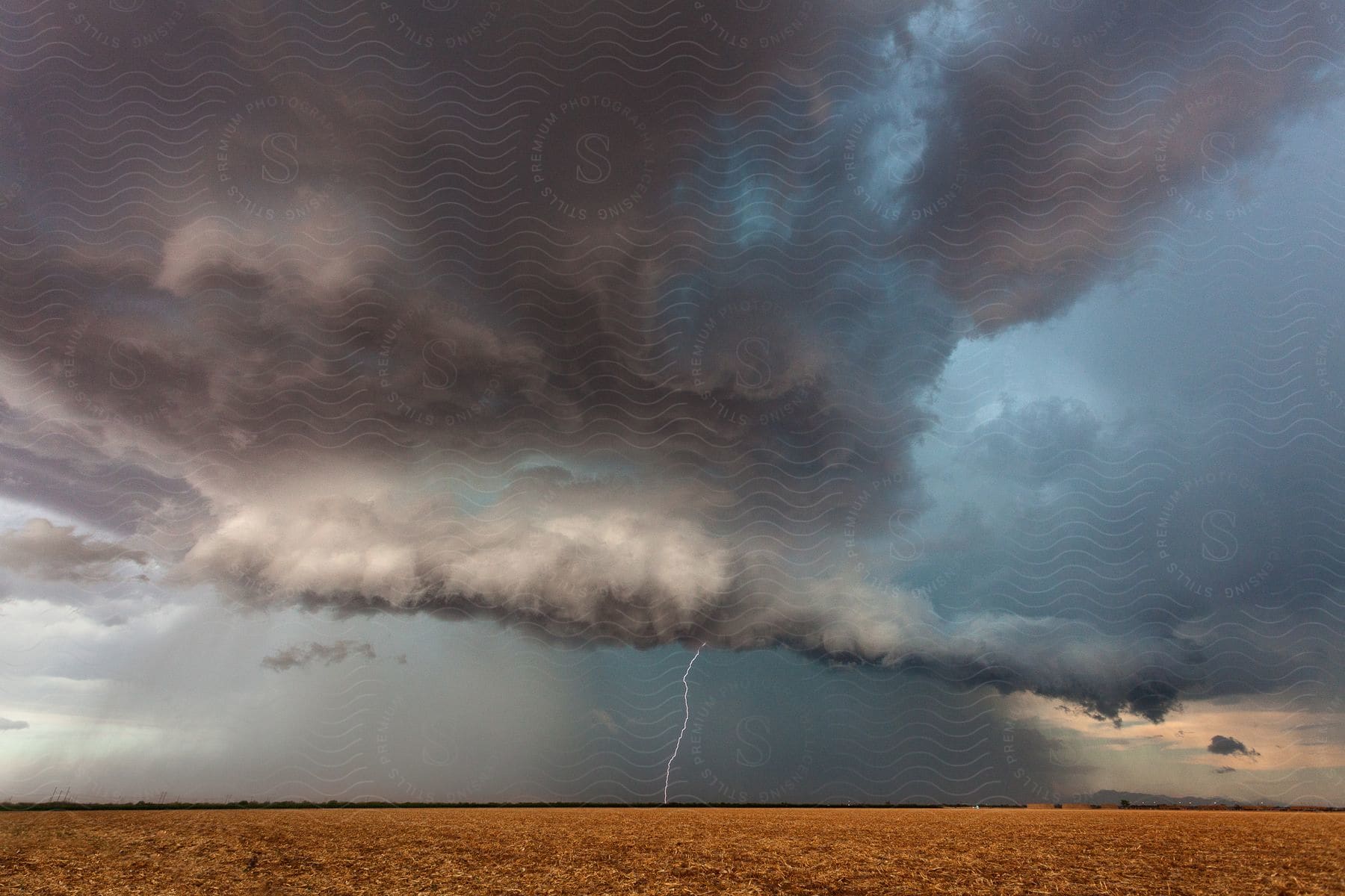 A lightning bolt strikes rural farmland from a dark storm cloud pouring rain on the land