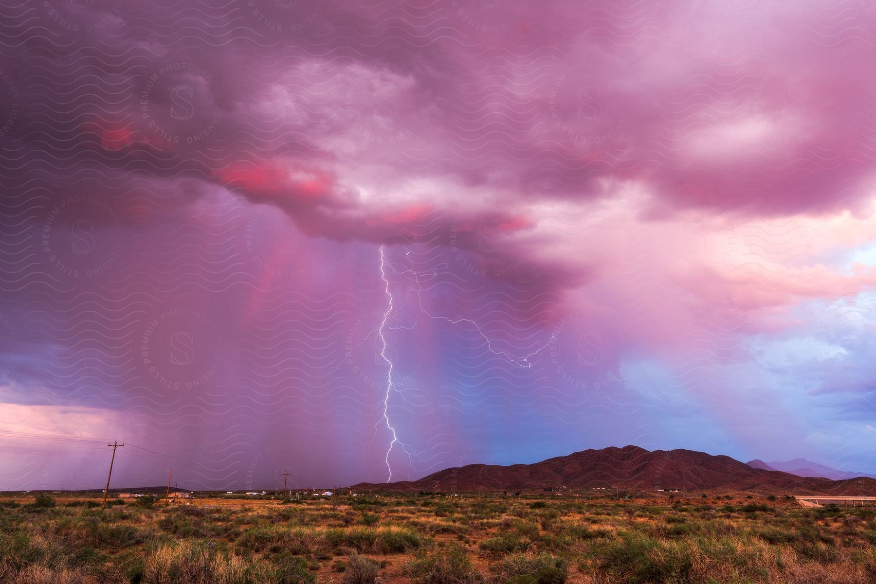 A small town under a pink cloudy sky with lightning illuminating the horizon above a green field near a majestic hill and an electric pole