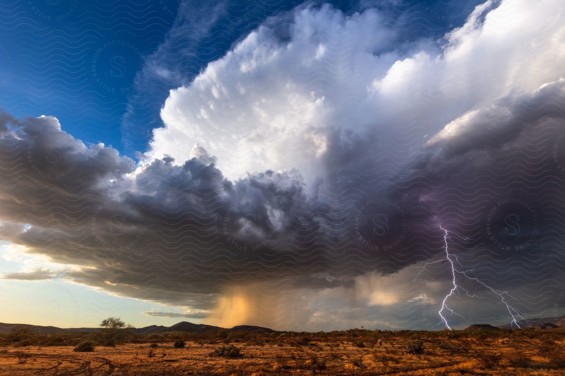 A dark supercell storm cloud pours down rain as lightning strikes near mountains in the distance