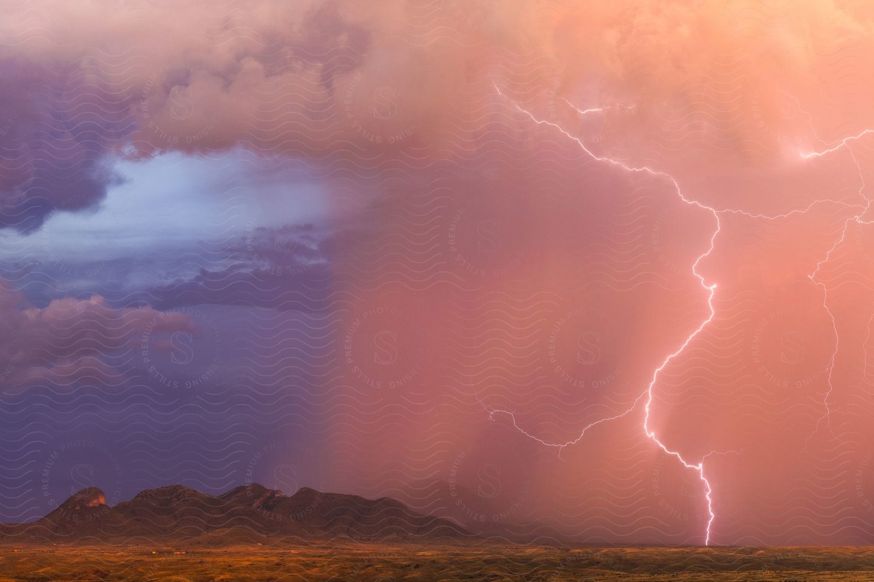 Rain falls from storm clouds over the mountains in the plains as lightning strikes along the lower slopes at sunset