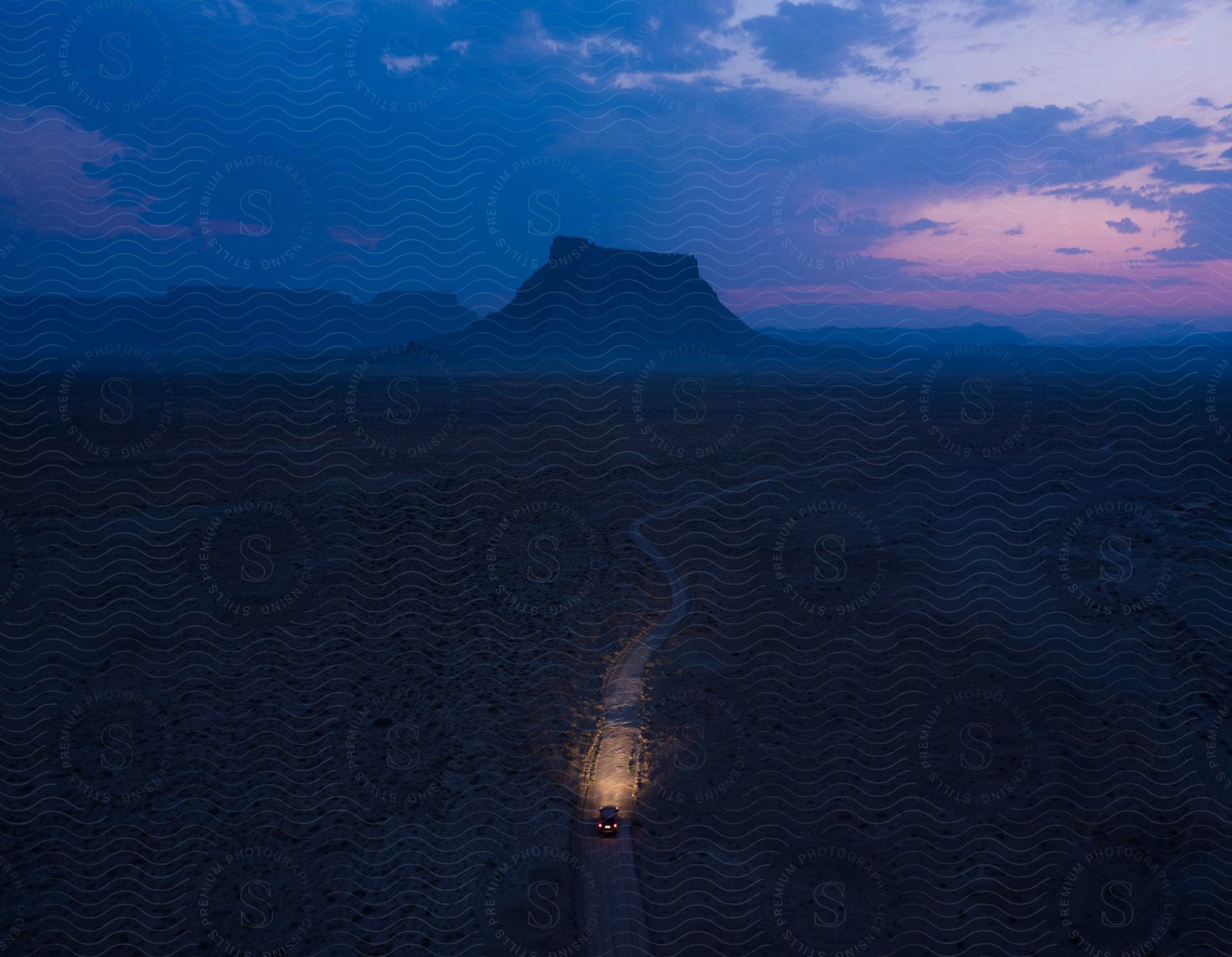 Automobile headlights glow as it travels on a road with buttes and mountains in the distance under a dark cloudy sky