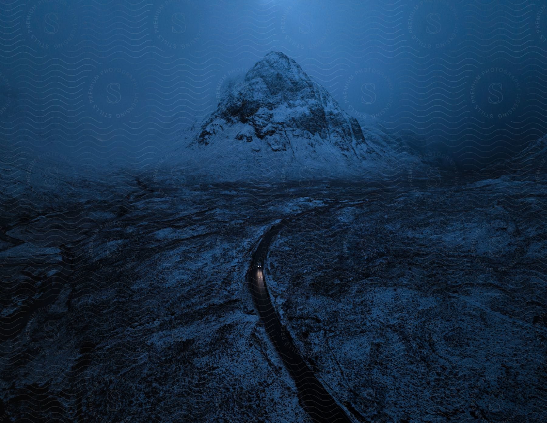 Automobile headlights glow on a road near a snow covered mountain under a dark hazy sky at night