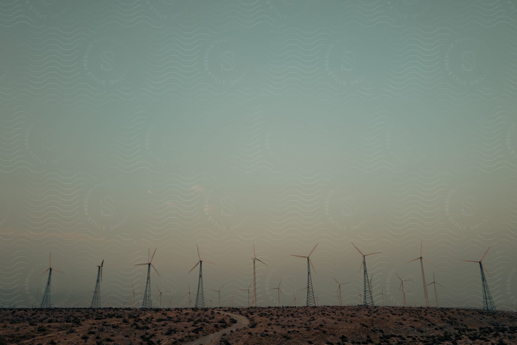 A road runs through the desert as wind turbines stretch across the landscape in a hazy sky