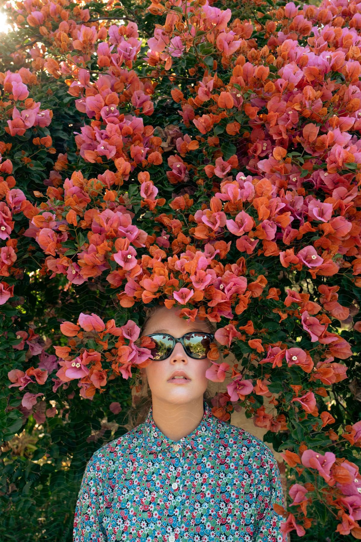 Woman wearing colorful shirt and sunglasses stands under bush filled with red flowers