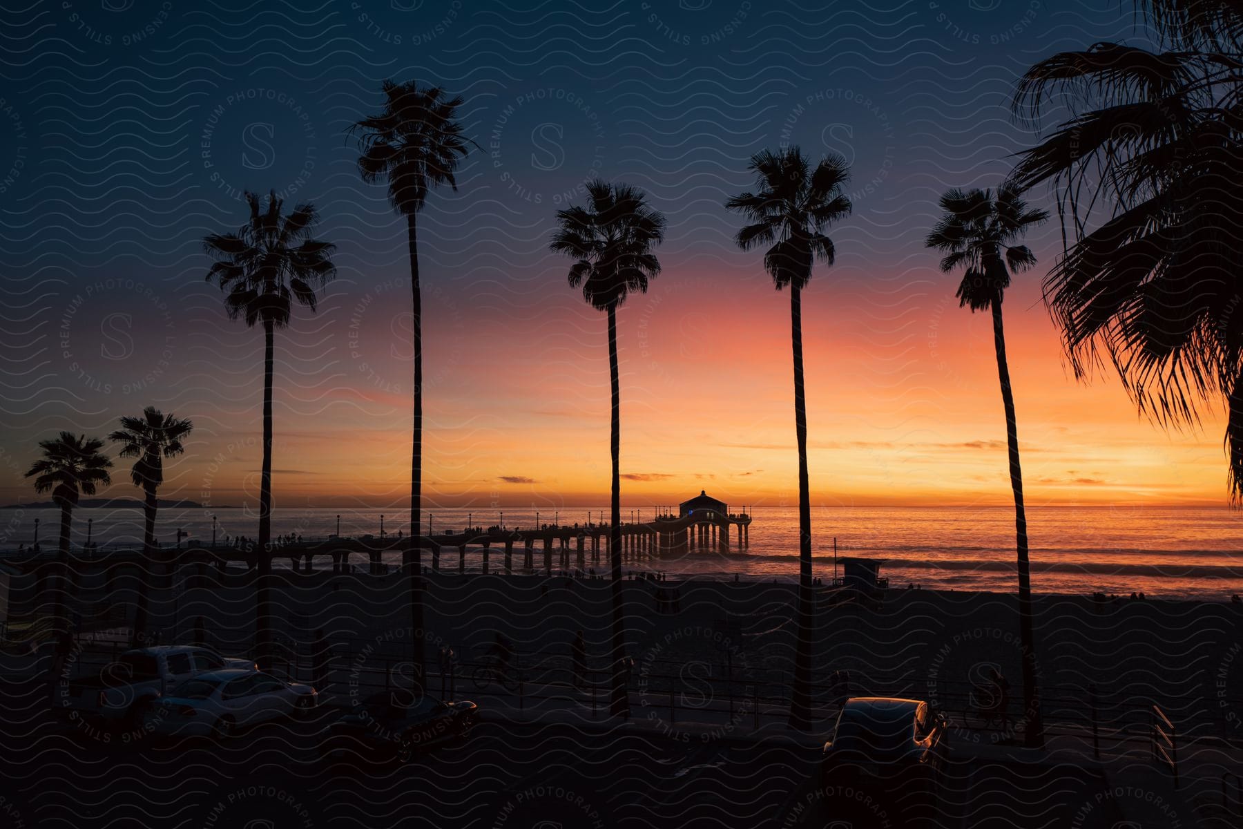 A pier leading out to calm waters at sunset with a few cars parked on the beach