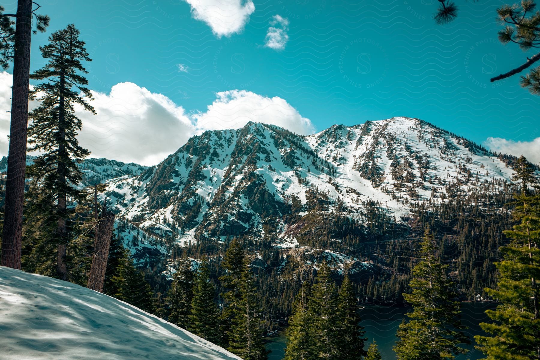 Snowcovered mountains stand majestically surrounded by trees and fluffy clouds in a natural landscape