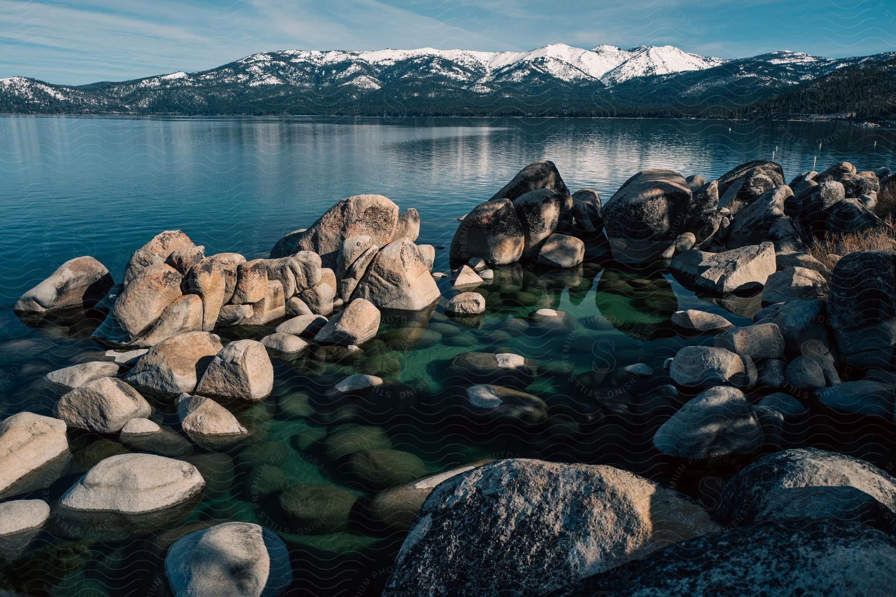 Rocks and boulders in water along the coast with sun shining on snowcovered mountains across the lake